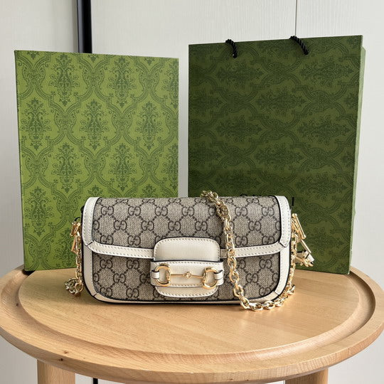 Gucci Horsebit 1955 Mini Bag in GG Supreme canvas with white leather trim and gold-tone chain strap, displayed on a wooden table with Gucci green gift boxes